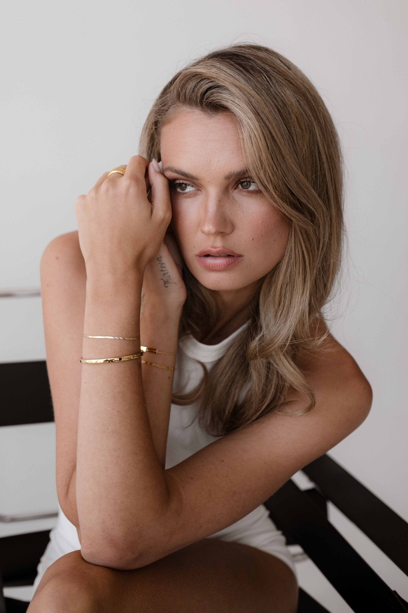 Blonde woman with wavy hair wearing gold bracelets and a white top, sitting on a chair, looking contemplative. Fashion, jewelry, beauty.
