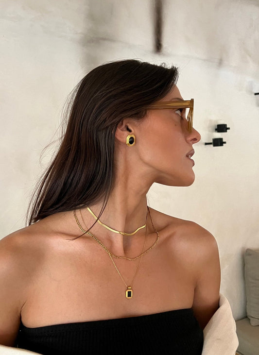 Woman wearing sunglasses, gold jewelry, and a black strapless top, looking sideways against a neutral background. Fashion accessories, elegant style.