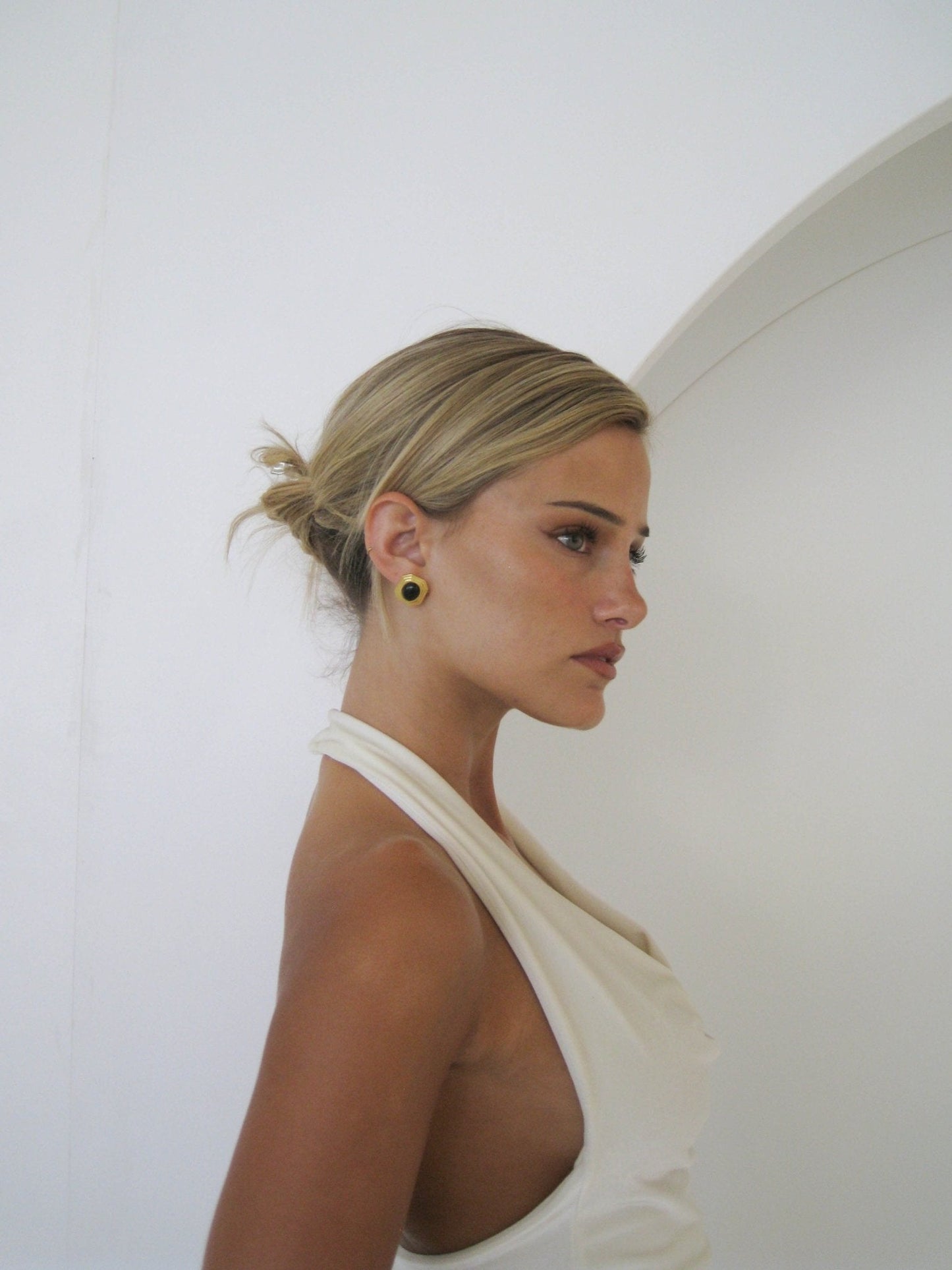 A woman in a white sleeveless top with a draped neckline, side profile, hair in a bun, standing against a white arched background.