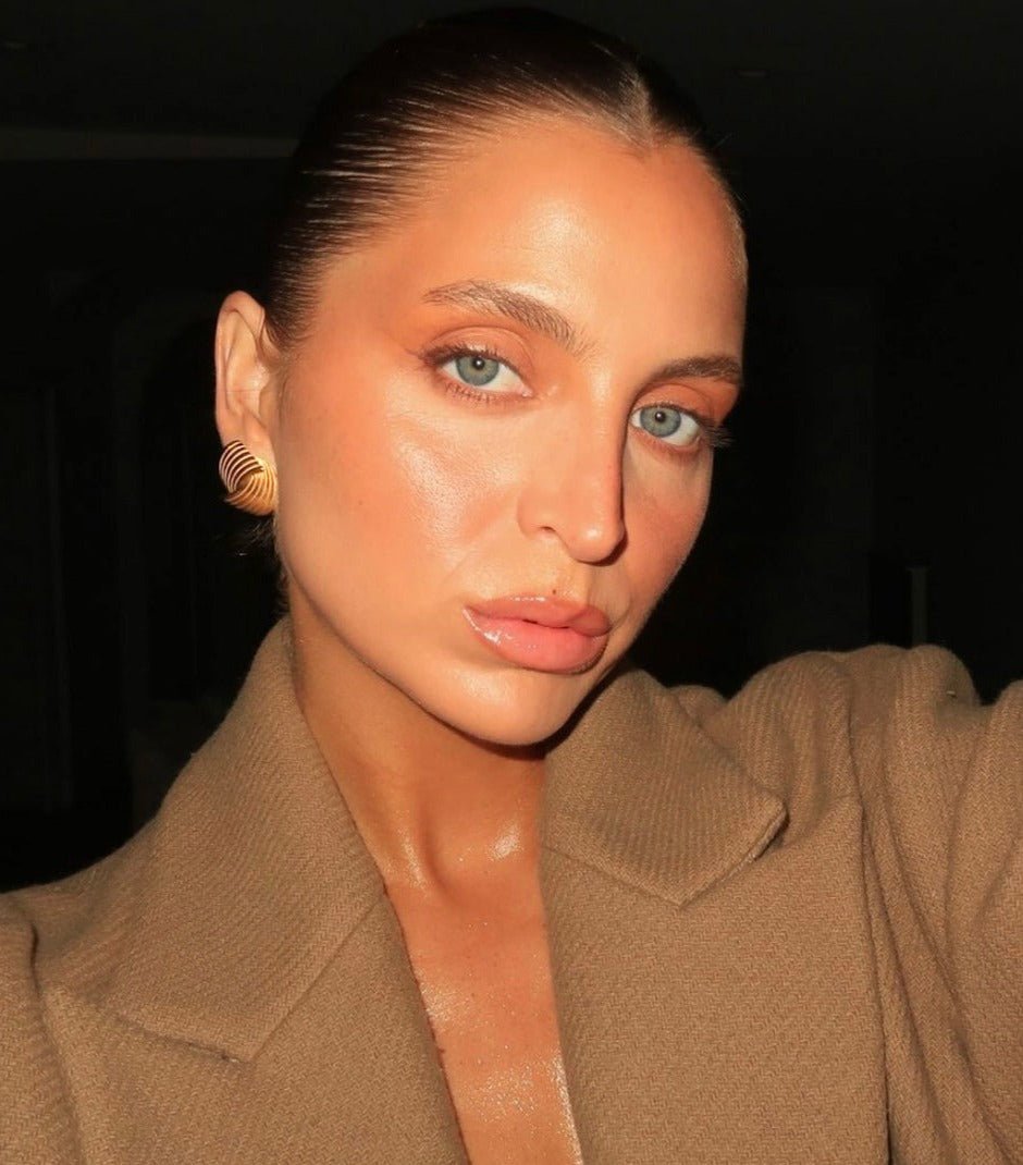 I don’t know who this person is, but the image shows a woman with slicked-back hair, wearing a brown coat and gold earrings, posing against a dark background.
