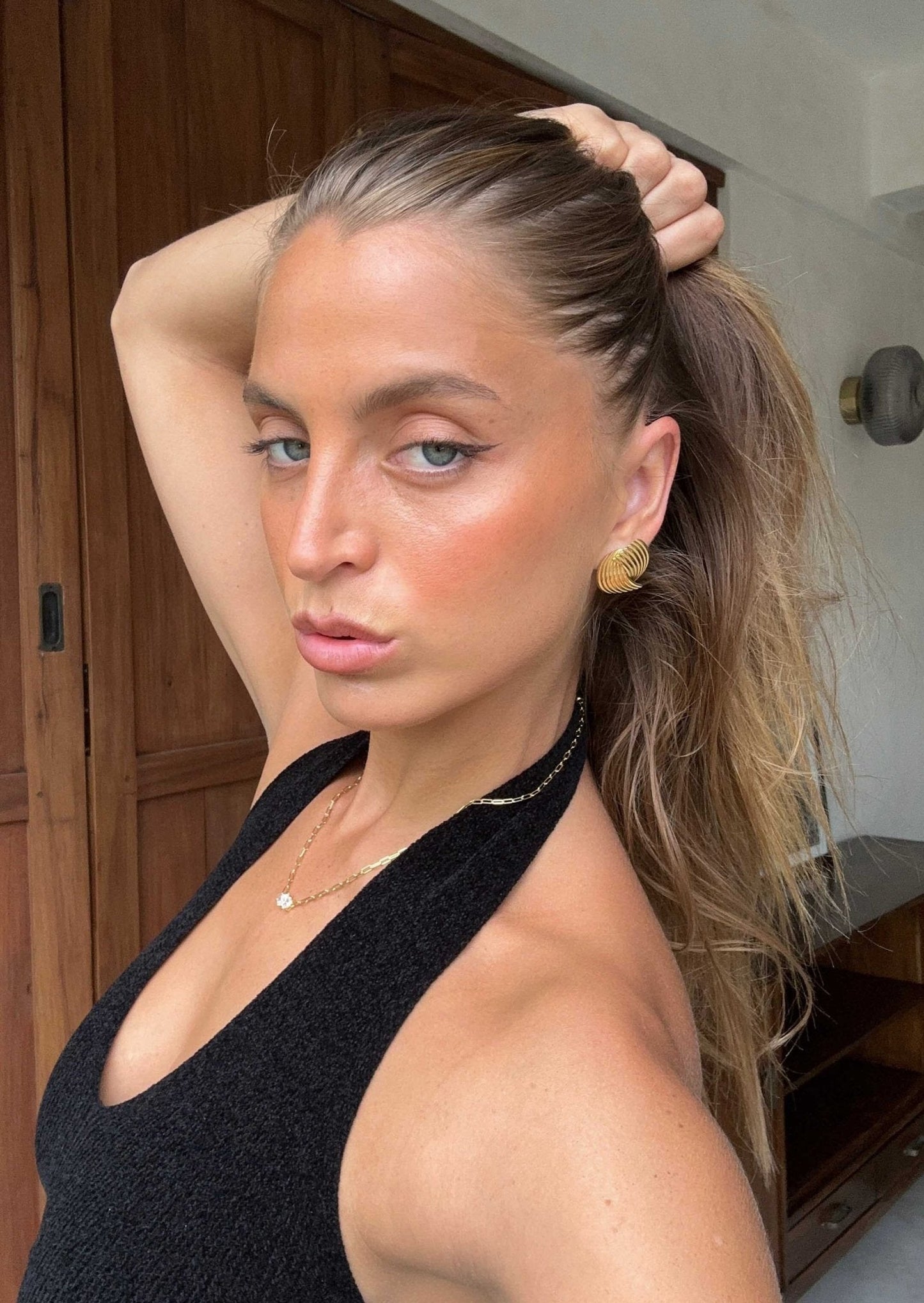 Woman with sleek ponytail and black halter top poses confidently indoors, showcasing gold earrings. Fashion, hairstyle, and jewelry style concept.