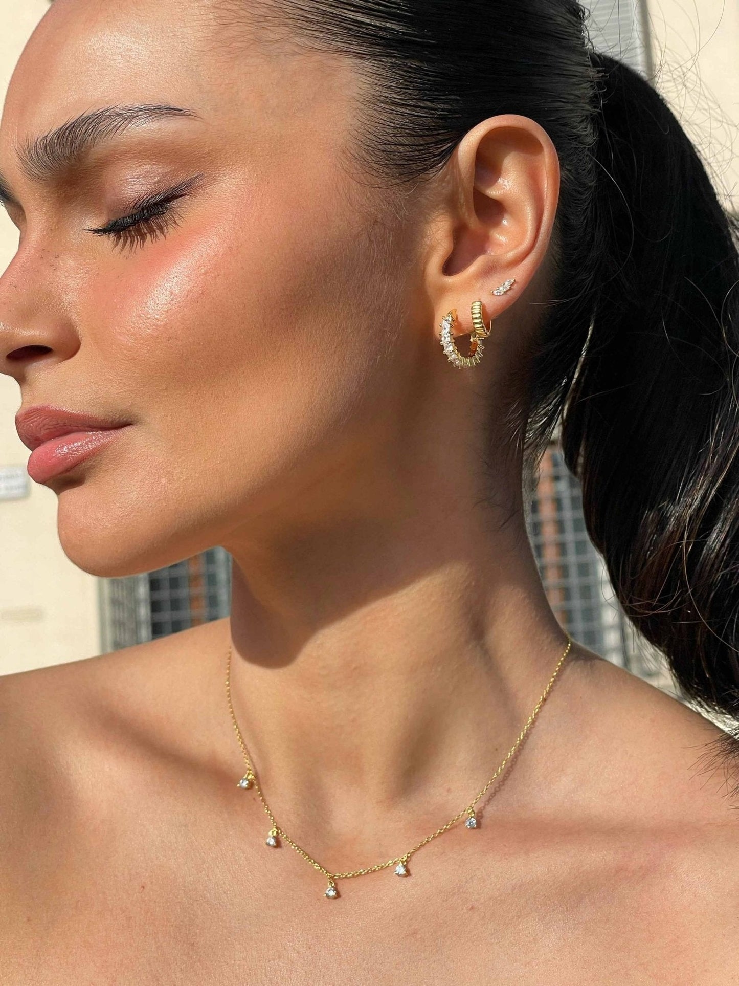 Woman wearing gold hoop earrings and a delicate gold necklace with small pendants, showcasing elegant jewelry fashion against a blurred urban background.