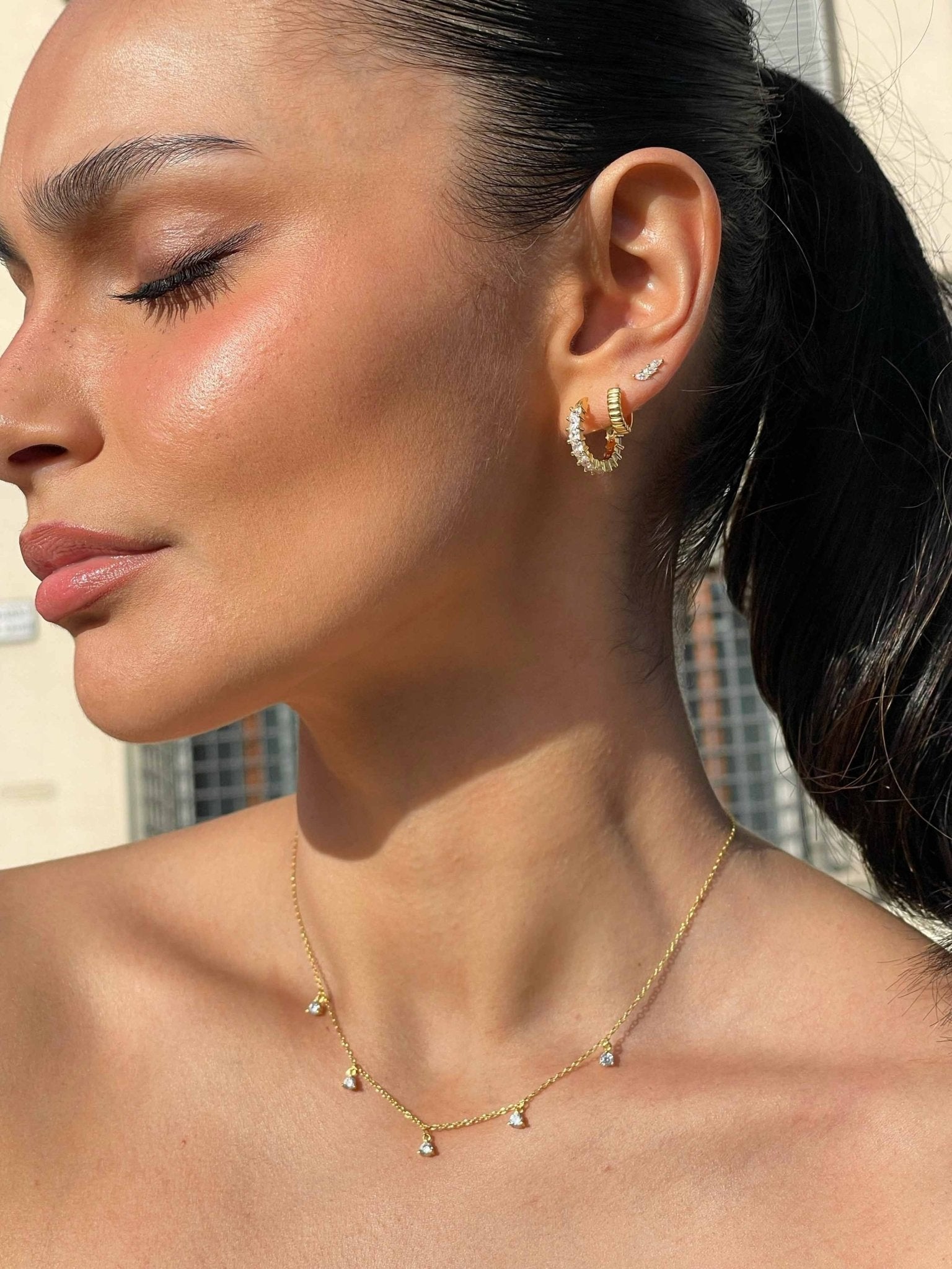 Woman wearing gold hoop earrings and a delicate gold necklace with small pendants, showcasing elegant jewelry fashion against a blurred urban background.