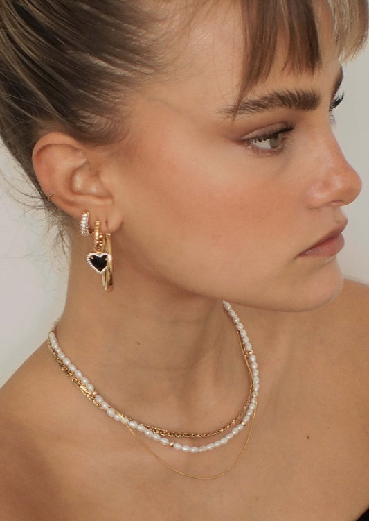Woman wearing layered gold and pearl necklaces, gold hoop earrings with a black heart charm, and a small hoop earring, showcasing elegant jewelry.