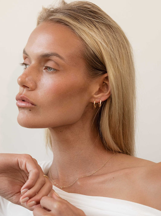 Blonde woman wearing minimalist gold hoop earrings and a delicate necklace, showcasing elegant jewelry fashion against a neutral background.