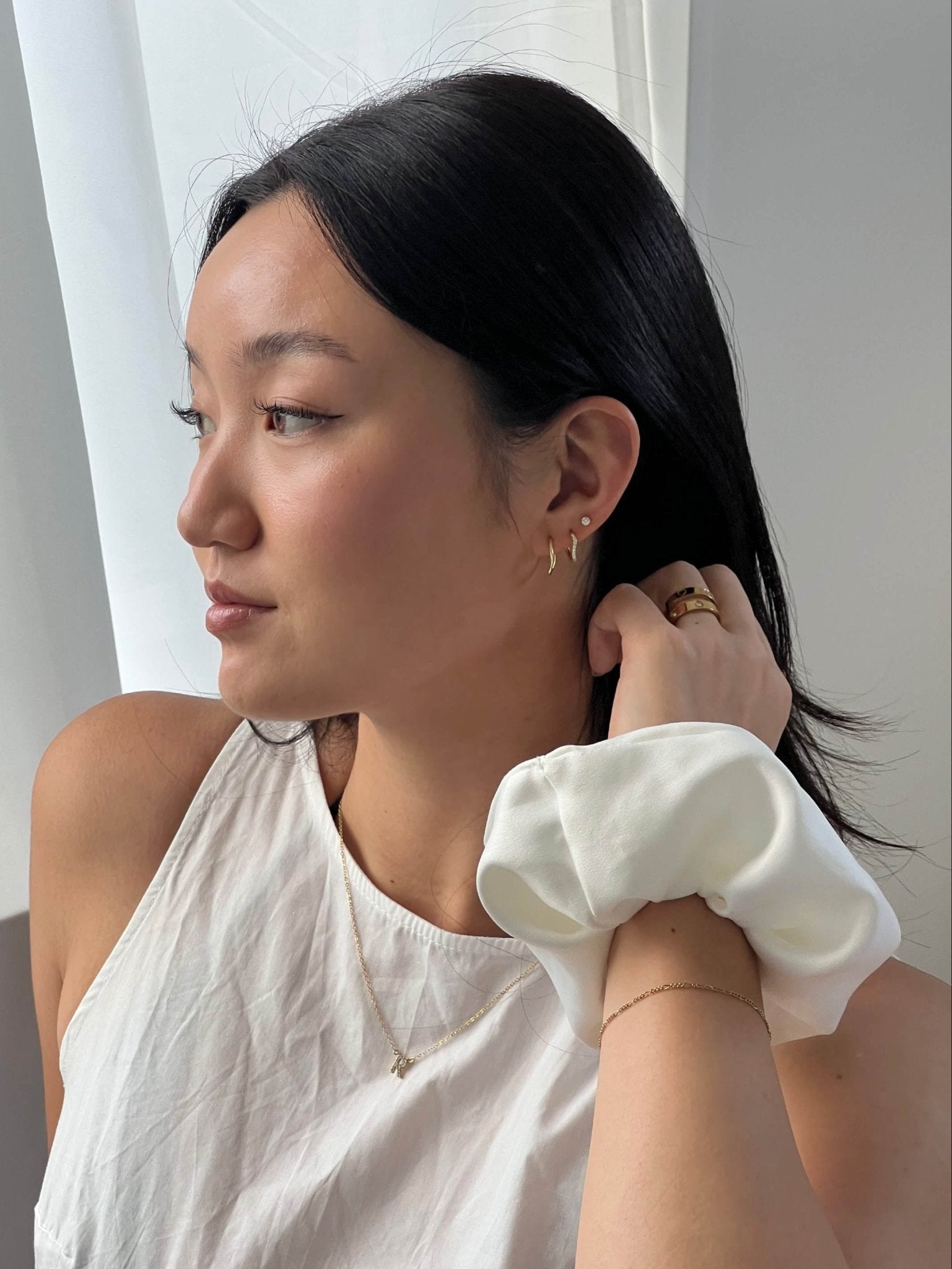 Woman with black hair wearing a white sleeveless top and gold jewelry, holding a white scrunchie. Natural light, minimalistic style.