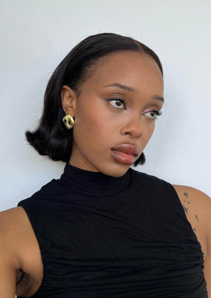 Young woman with short black hair, wearing gold hoop earrings and a sleeveless black top, gazes to the side against a plain white background.