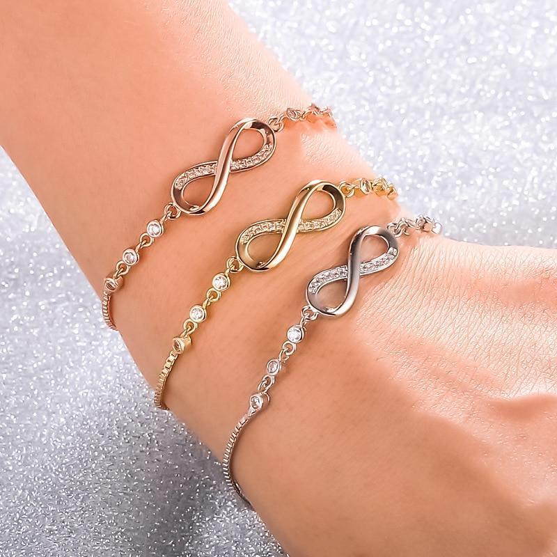 Three infinity symbol bracelets in gold, silver, and rose gold on a wrist, adorned with sparkling crystals, against a glittery background.