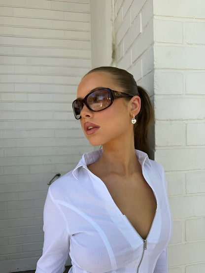 Woman wearing sunglasses and a white shirt against a light-colored wall.