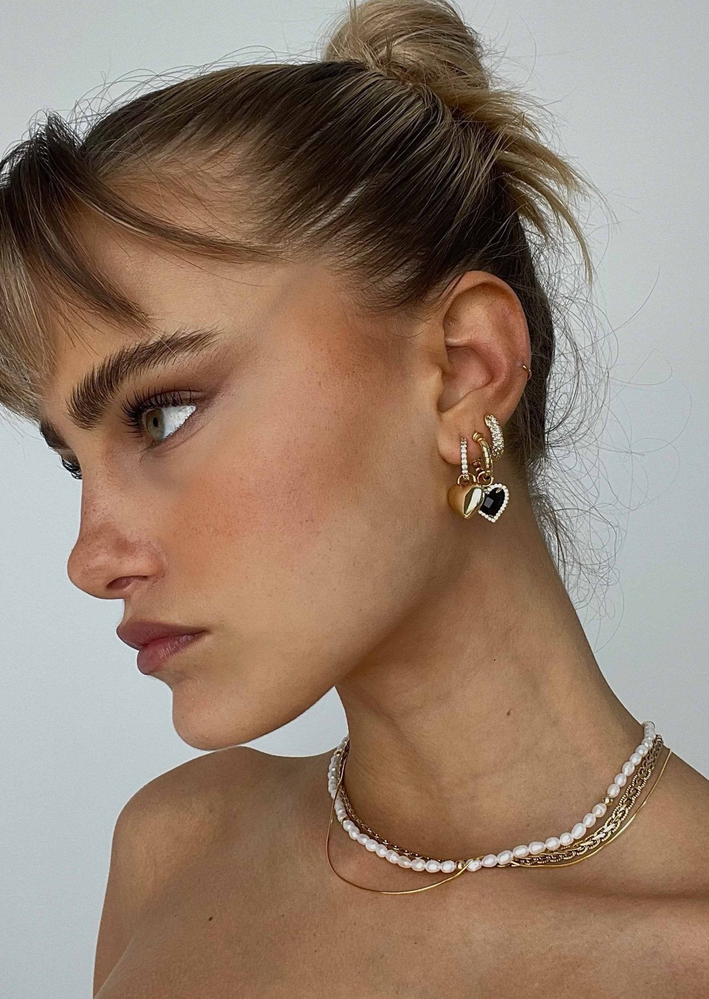 Close-up of a person wearing gold hoop earrings and pearl necklaces against a neutral background