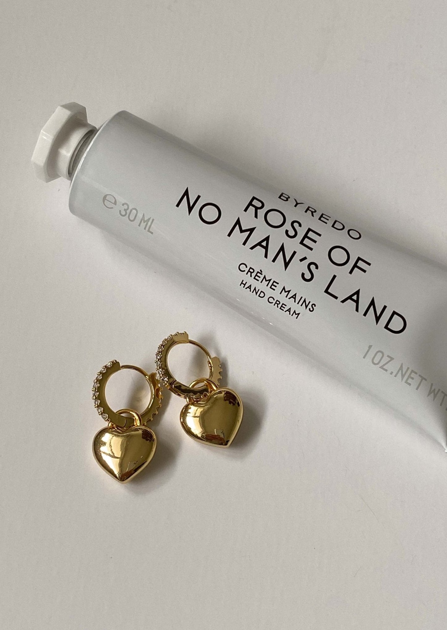 Byredo 'Rose of No Man's Land' hand cream with gold heart-shaped earrings on a light gray background
