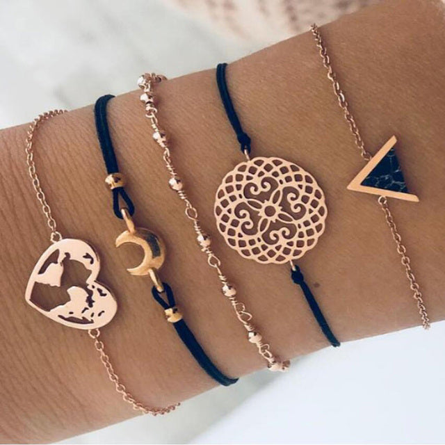 Five stylish bracelets on wrist featuring rose gold and black designs, including a world map, crescent moon, and geometric patterns. Fashion accessories.