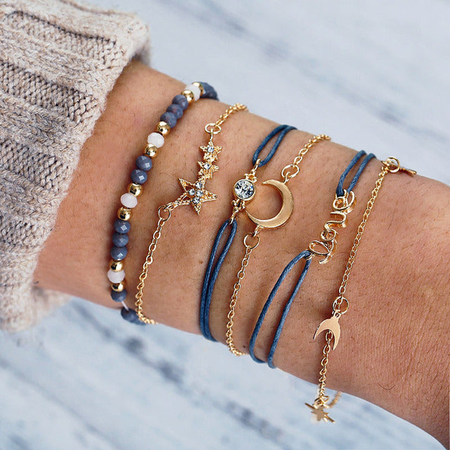 A wrist adorned with layered gold and blue bracelets featuring star, moon, and bead designs on a cozy sweater background. Fashionable jewelry accessories.
