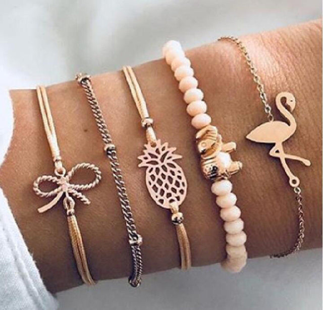 Four stylish gold and pink bracelets on wrist, featuring bow, pineapple, elephant, and flamingo charms, with beads and chain details. Fashion accessories.