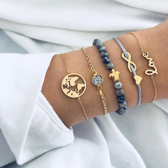 Gold and silver layered bracelets on wrist, featuring world map charm, infinity symbol, and beaded design. Fashionable jewelry accessories.