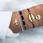 A wrist adorned with trendy gold and black bracelets featuring a plane charm, cowrie shell, and world map design. Fashionable jewelry accessories.
