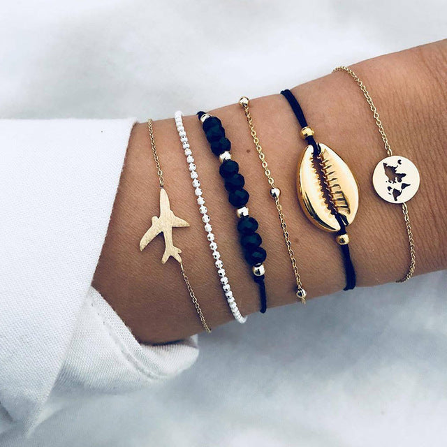 A wrist adorned with trendy gold and black bracelets featuring a plane charm, cowrie shell, and world map design. Fashionable jewelry accessories.