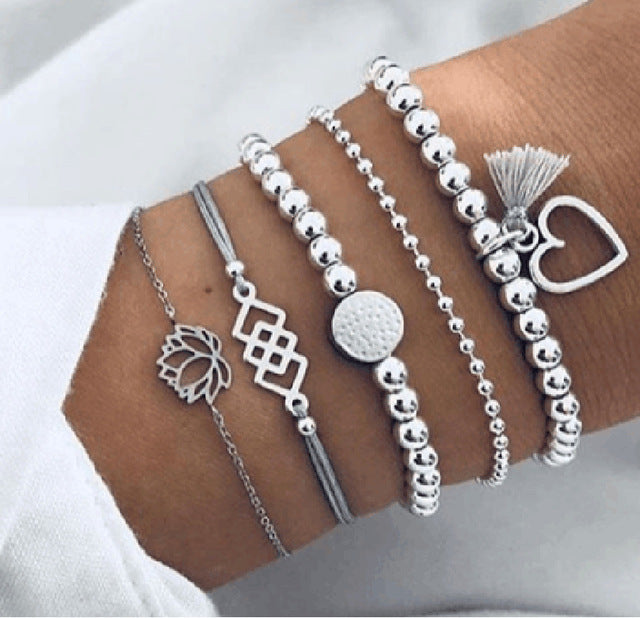 Silver bracelet stack on wrist featuring beaded, heart charm, lotus, and geometric designs; trendy women's fashion jewelry accessories.