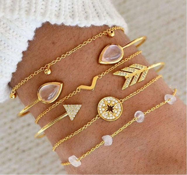 Gold layered bracelets with geometric charms and gemstones on a wrist, featuring arrow, star, and teardrop designs. Fashionable jewelry accessory.