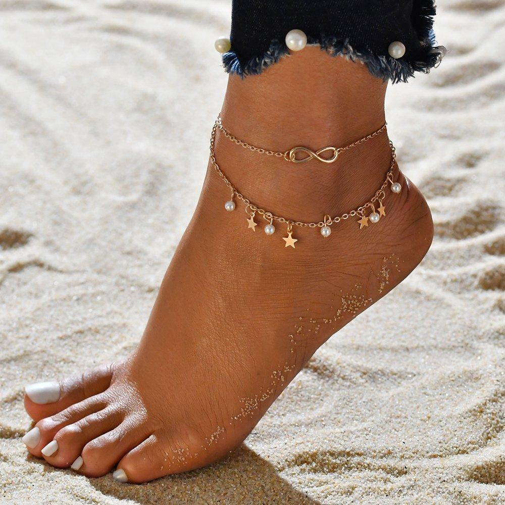 Bare foot on sand wearing a gold anklet with infinity symbol, star charms, and pearls; beach jewelry, summer fashion accessory, boho style.