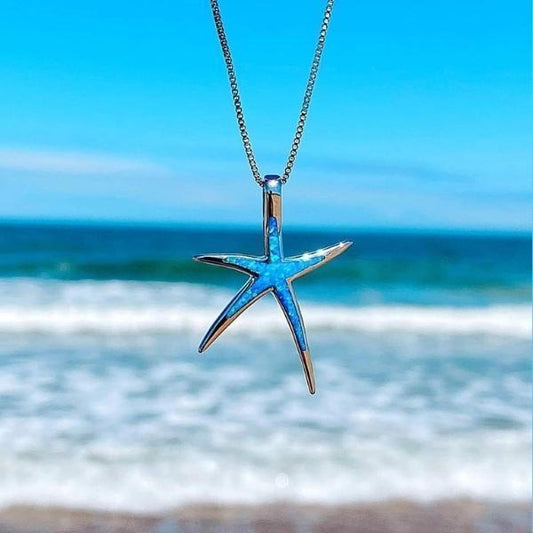 Starfish pendant necklace with blue opal inlay, hanging against a vibrant ocean backdrop. Perfect beach jewelry, coastal fashion accessory.