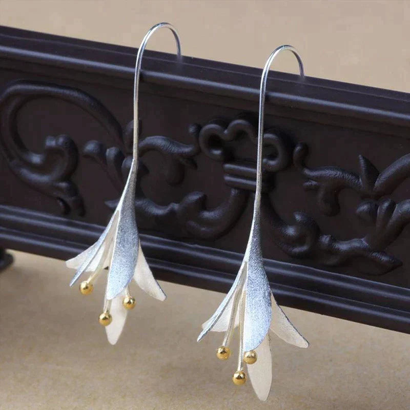 Elegant silver drop earrings with floral design and gold bead accents, displayed on ornate dark wood background. Fashion jewelry accessory.