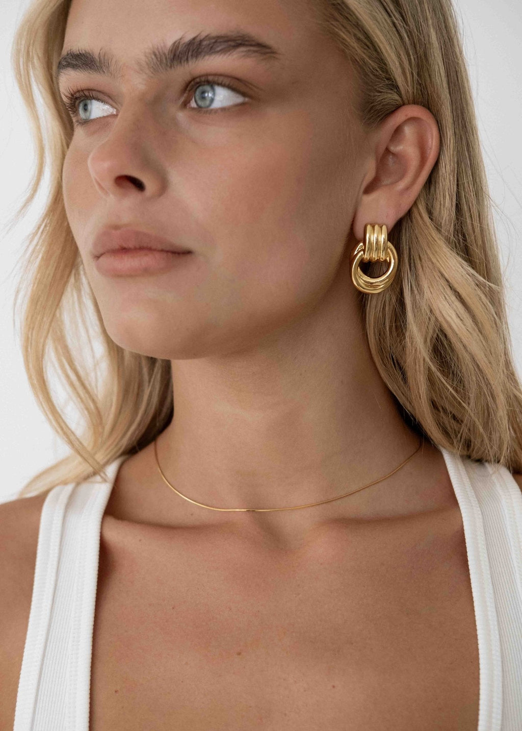 Woman wearing gold hoop earrings and a thin gold necklace, looking to the side. Blonde hair, white tank top, minimalistic jewelry fashion.
