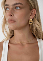 Woman wearing gold hoop earrings and a thin gold necklace, looking to the side. Blonde hair, white tank top, minimalistic jewelry fashion.