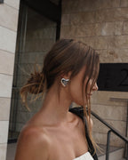 Woman with messy bun hairstyle wearing large silver heart-shaped earrings, standing against a modern stone building background. Fashion accessory.