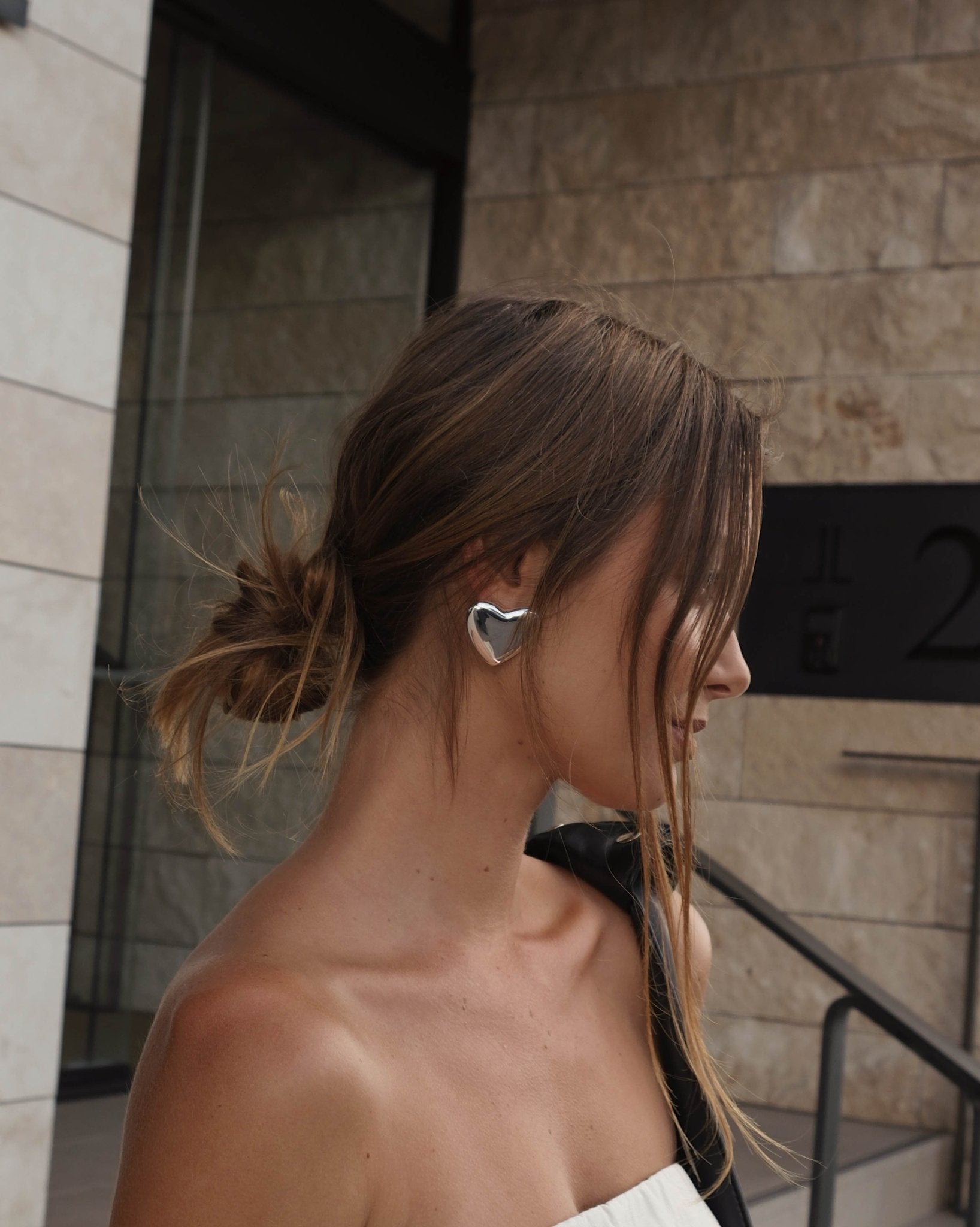 Woman with messy bun hairstyle wearing large silver heart-shaped earrings, standing against a modern stone building background. Fashion accessory.