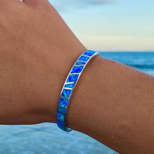 Hand wearing a blue opal inlay bracelet by the ocean, showcasing vibrant colors and silver accents, perfect for beach jewelry fashion enthusiasts.