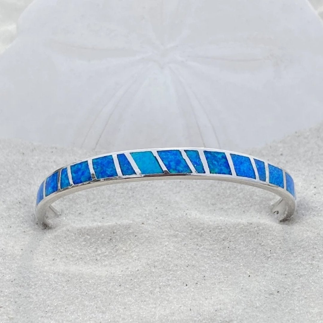 Silver bracelet with blue opal inlay on sand, featuring geometric design. Elegant jewelry piece, perfect for beach-themed fashion and accessories.
