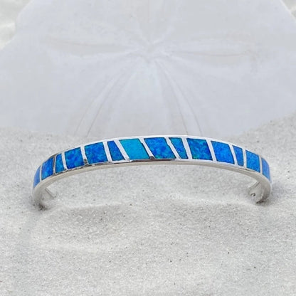 Silver bracelet with blue opal inlay on sand, featuring geometric design. Elegant jewelry piece, perfect for beach-themed fashion and accessories.