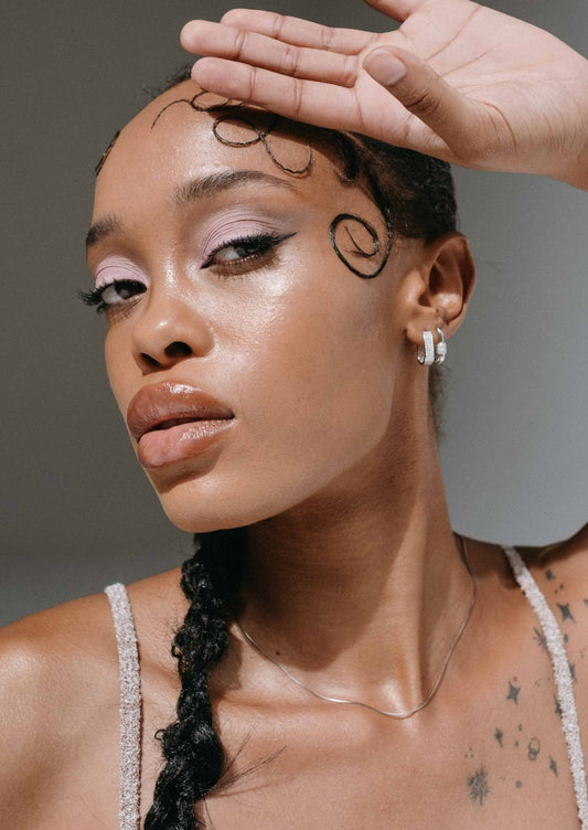 Woman with braided hair and star tattoos, wearing silver hoop earrings, poses with hand on forehead. Elegant makeup, glossy lips, and artistic hairstyle.