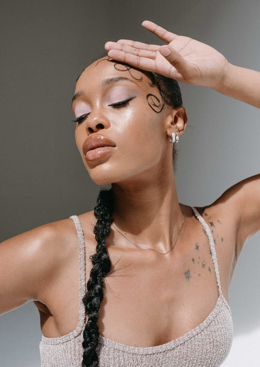 Woman with braided hair and tattoos poses in a sleeveless top, showcasing elegant makeup and jewelry. Fashion photography, beauty portrait.