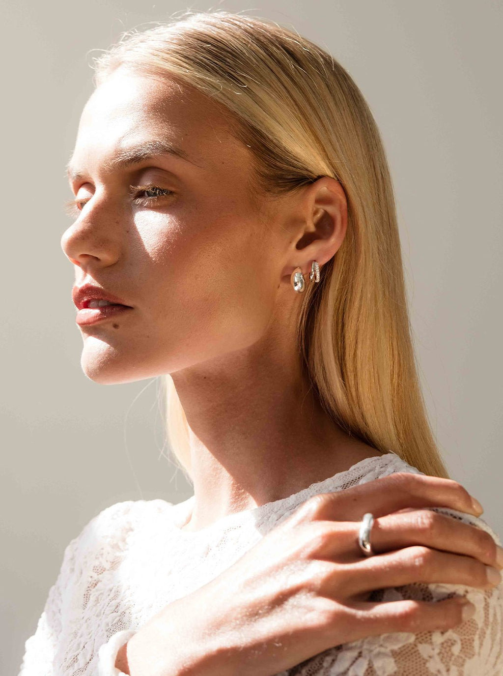 A woman with long blonde hair wearing silver hoop earrings and a white lace top, posing in soft natural light, showcasing elegant jewelry and fashion.