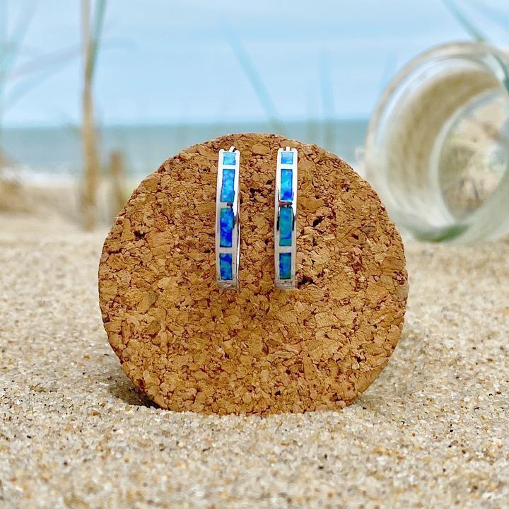 Silver hoop earrings with blue opal inlay displayed on a cork stand, set against a sandy beach background with a glass bottle.