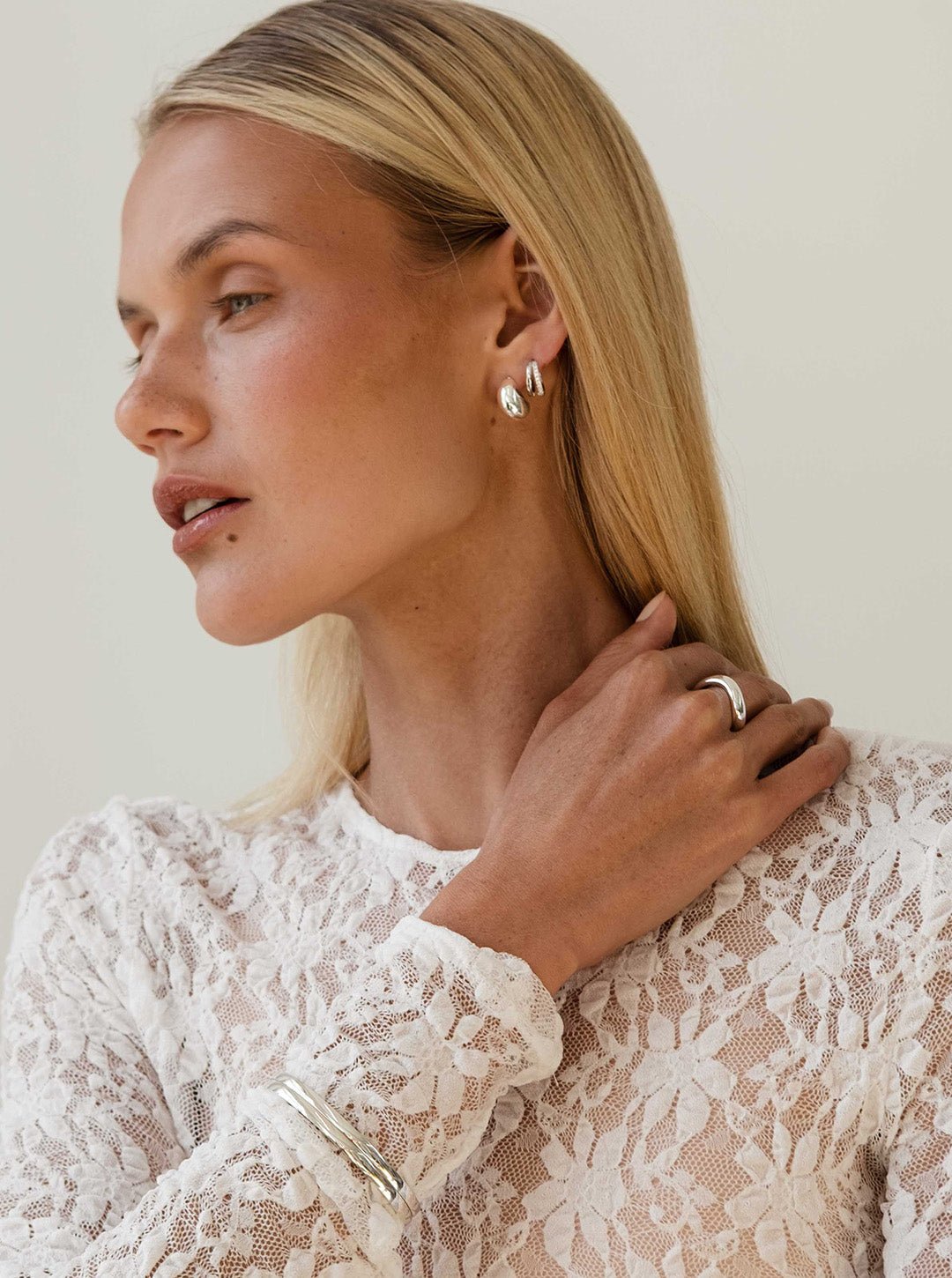 Woman wearing elegant silver jewelry, including hoop earrings, a ring, and bracelets, paired with a white lace top. Fashion accessories.