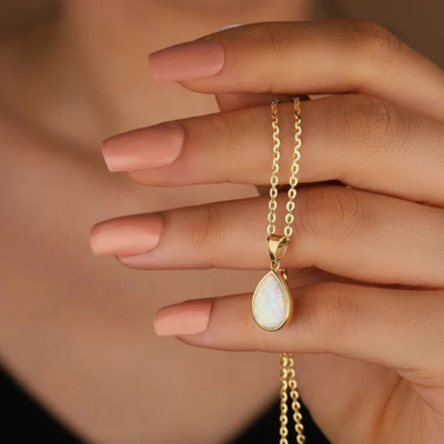 Gold chain necklace with teardrop opal pendant held by hand with manicured nails, elegant jewelry, fashion accessory, close-up.