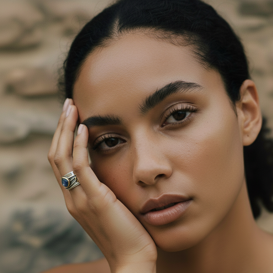 Close-up of a woman with curly hair, wearing a blue gemstone ring, resting her face on her hand against a textured stone background.