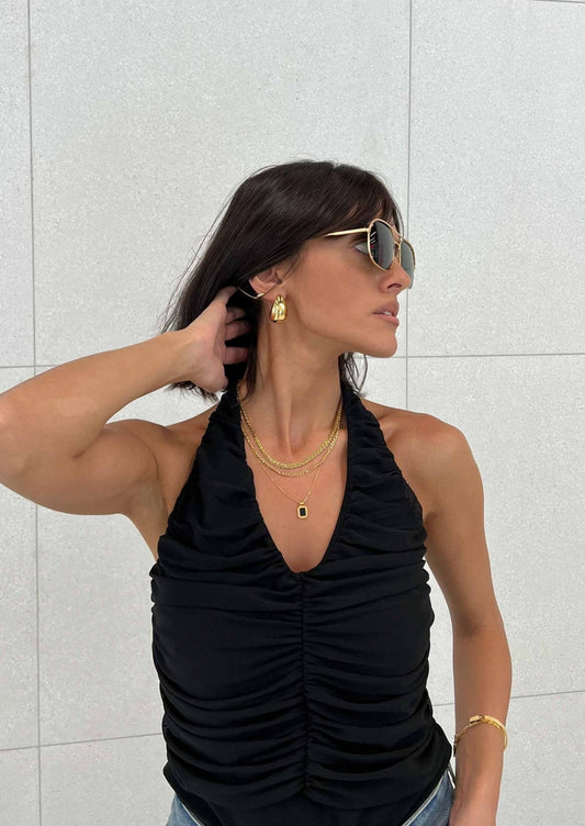 Woman in stylish black ruched halter top, gold layered necklaces, and sunglasses, posing against a light gray background. Fashionable jewelry and attire.