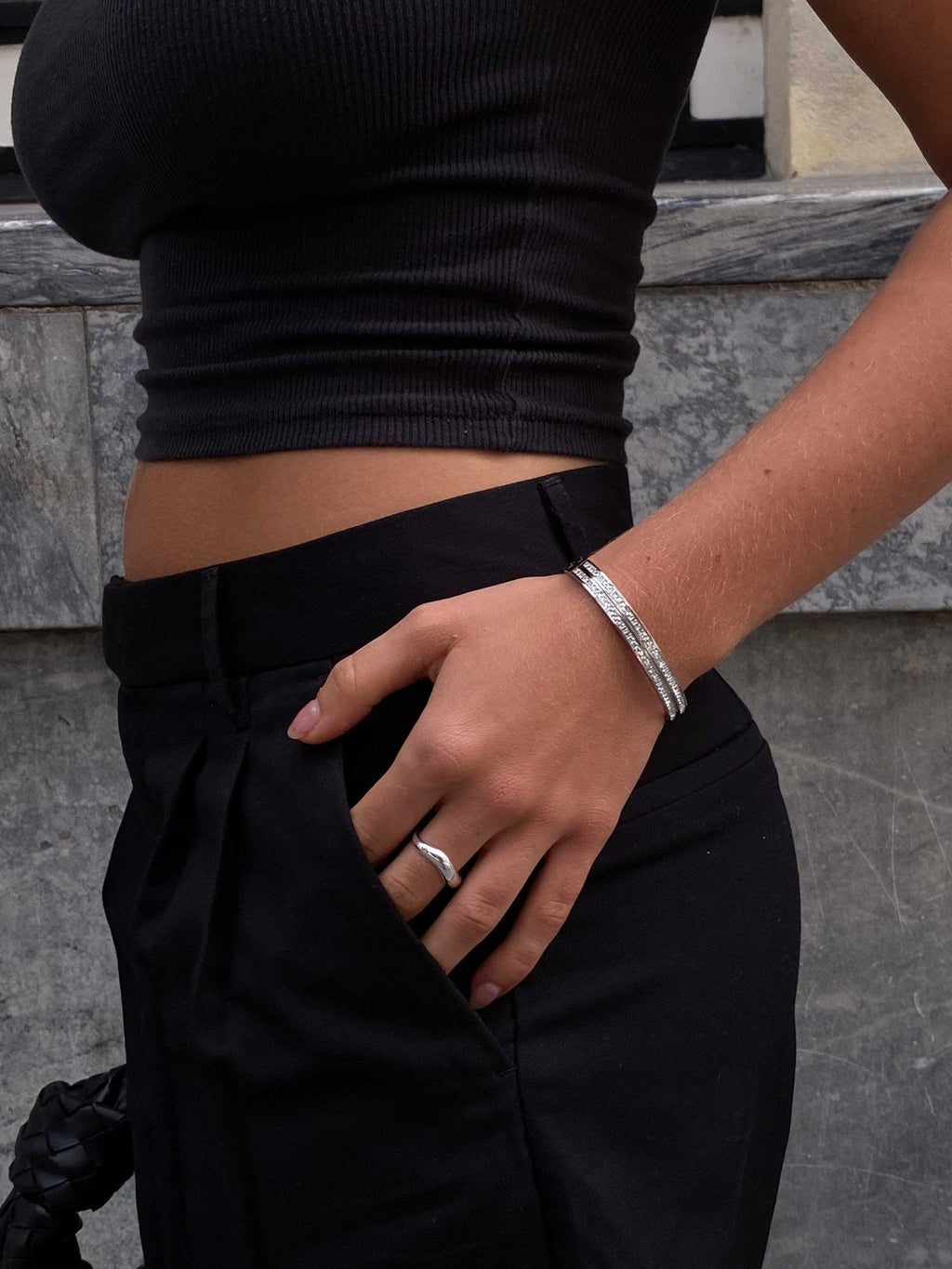 Woman in black crop top and pants, hand in pocket, wearing silver bracelet and ring. Fashionable street style, minimalist jewelry, urban background.