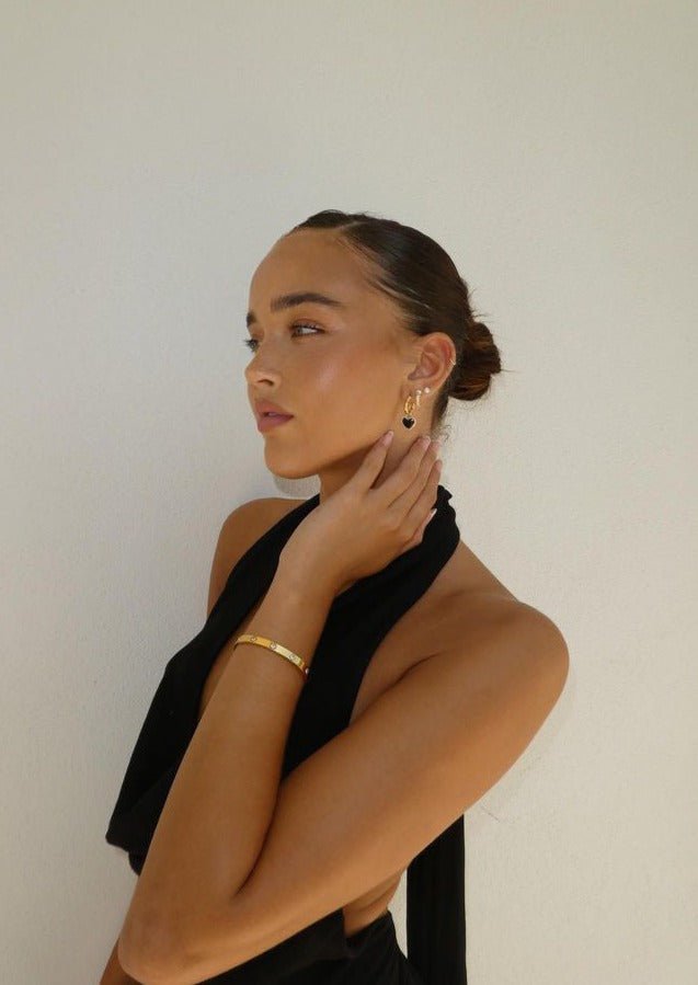 Young woman in elegant black dress, gold jewelry, sleek bun hairstyle, posing against a plain white background. Fashion, style, elegance.
