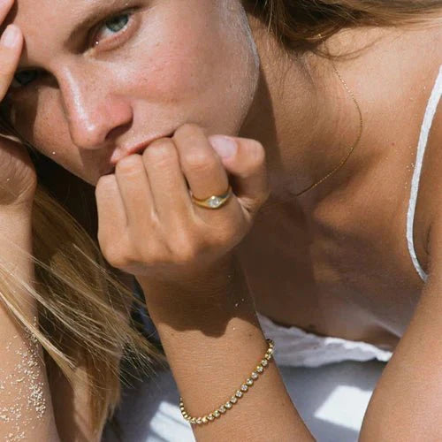 Woman wearing gold jewelry, including a delicate bracelet and ring, with sandy skin, gazing thoughtfully. Fashion, beach, summer accessories.
