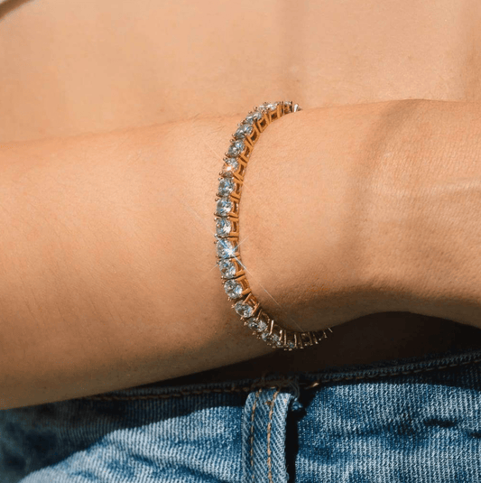 Gold tennis bracelet with sparkling diamonds on a woman's wrist, paired with blue denim jeans. Elegant jewelry, luxury fashion accessory.