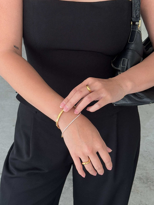 Woman in black outfit showcasing gold bracelet, pearl bracelet, and rings, holding a black leather bag. Fashion accessories, elegant style.
