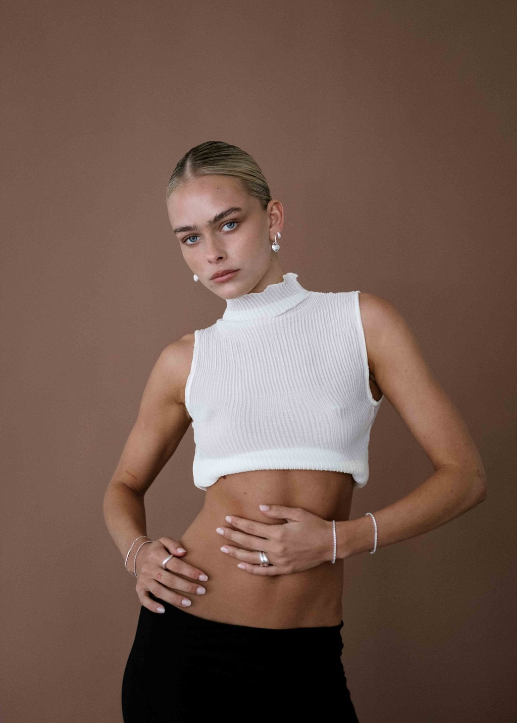 Woman in white sleeveless turtleneck and black pants posing against brown background, showcasing minimalist fashion style and pearl earrings.