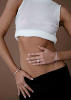 Woman in white ribbed crop top and black skirt, showcasing silver rings and bracelets on hands, against a brown background. Fashion jewelry.