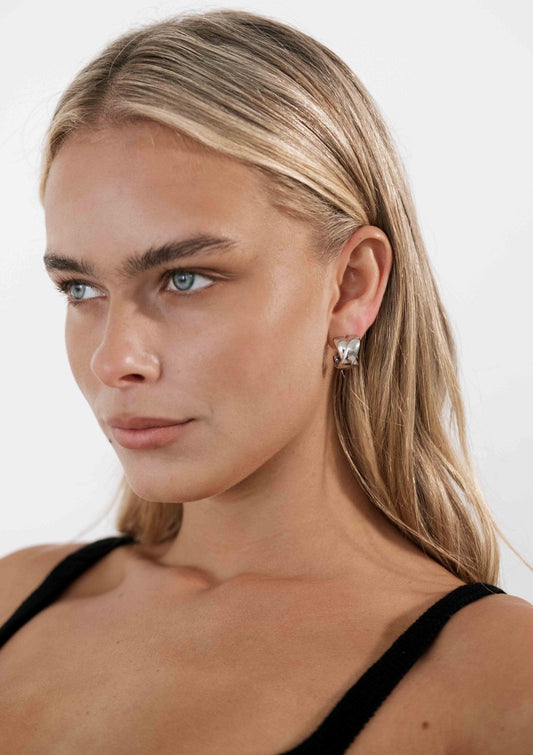 Blonde woman wearing silver hoop earrings, black top, and natural makeup, gazing to the side against a plain white background. Fashion jewelry portrait.