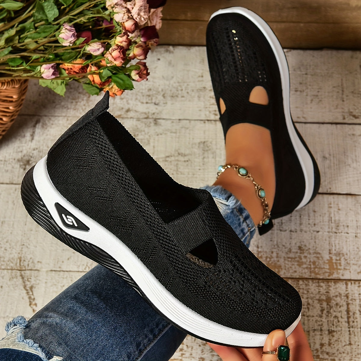 Black slip-on shoes with white soles worn by a person on a wooden floor.